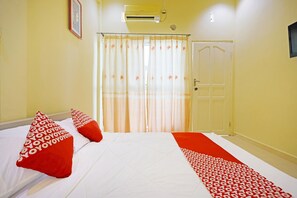 Habitación de hotel con cama, almohadones rojos, cubrecama blanco, corredor rojo y blanco estampado, puerta blanca y ventana con cortinas.