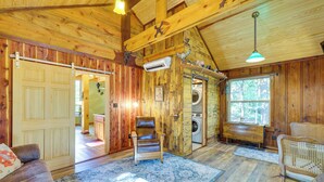 A wooden cabin interior with a sofa, chairs, a rug, a washing machine, and a large window. - Pet-Friendly Eureka Springs Rental w/ Private Deck (Eureka Springs)