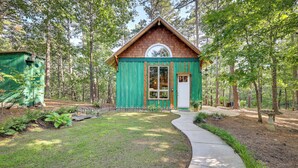 A small, rustic cabin with a green exterior and a white door, surrounded by a forested area with a paved path leading to it. - Pet-Friendly Eureka Springs Rental w/ Private Deck (Eureka Springs)