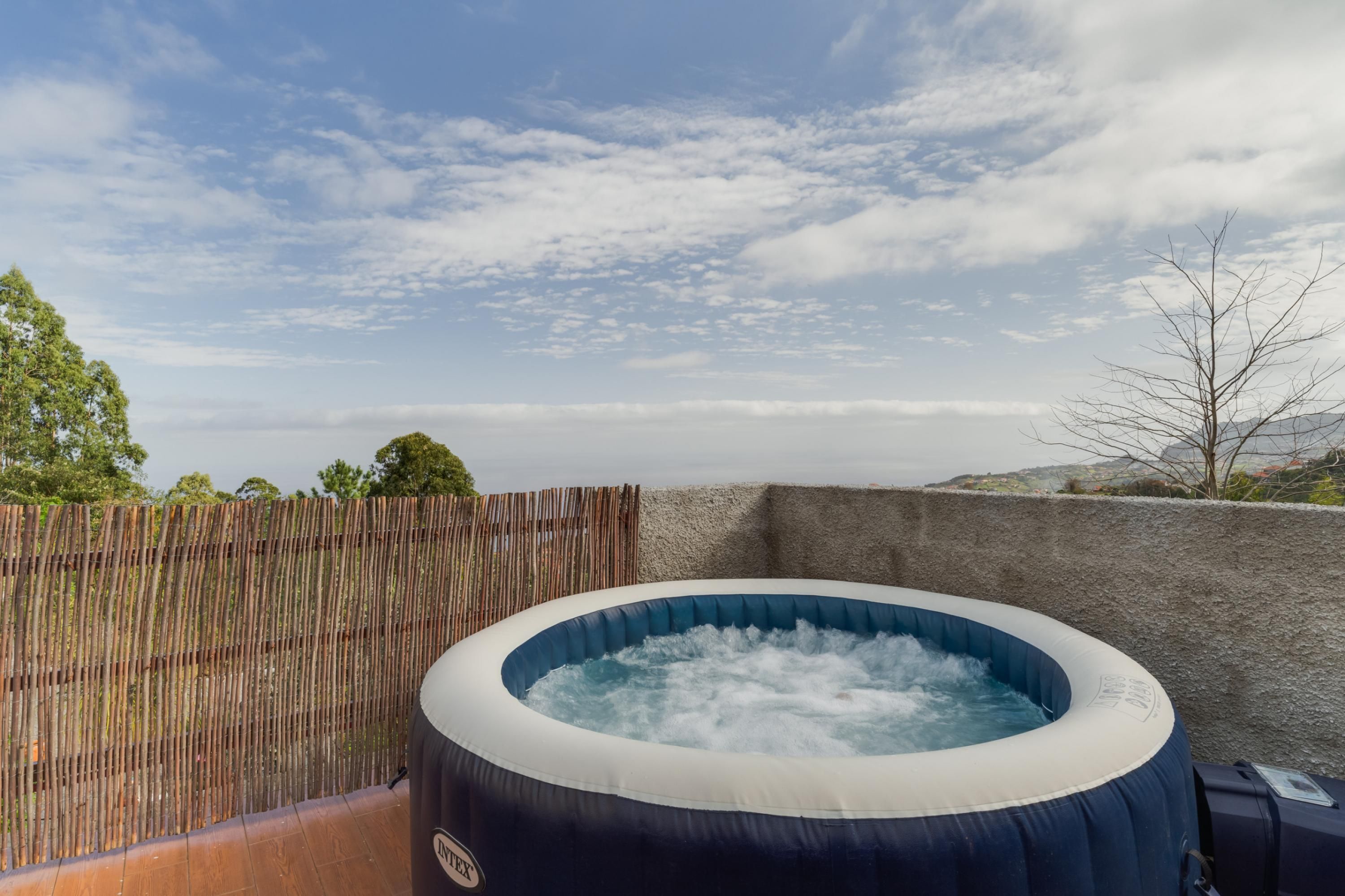 An inflatable hot tub with a visible brand logo, surrounded by a wooden fence and a stone wall, set against a backdrop of a clear sky and distant landscape.