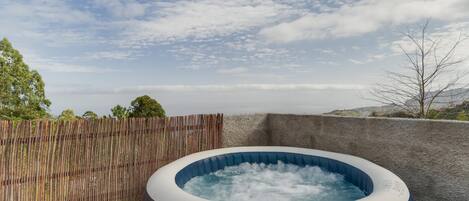 An inflatable hot tub with a visible brand logo, surrounded by a wooden fence and a stone wall, set against a backdrop of a clear sky and distant landscape.