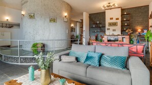 Una sala de estar moderna con un sofá gris, una mesa de centro y una chimenea. Se ve una escalera que sube a un nivel superior, y un área de cocina al fondo.