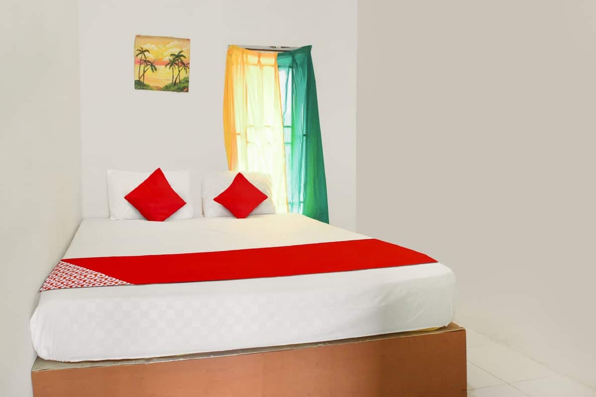 Ein Bett mit weißer Matratze, roten und weißen Kissen und einer roten Decke. An der Wand hängt ein Gemälde und ein Fenster mit grünen Vorhängen.