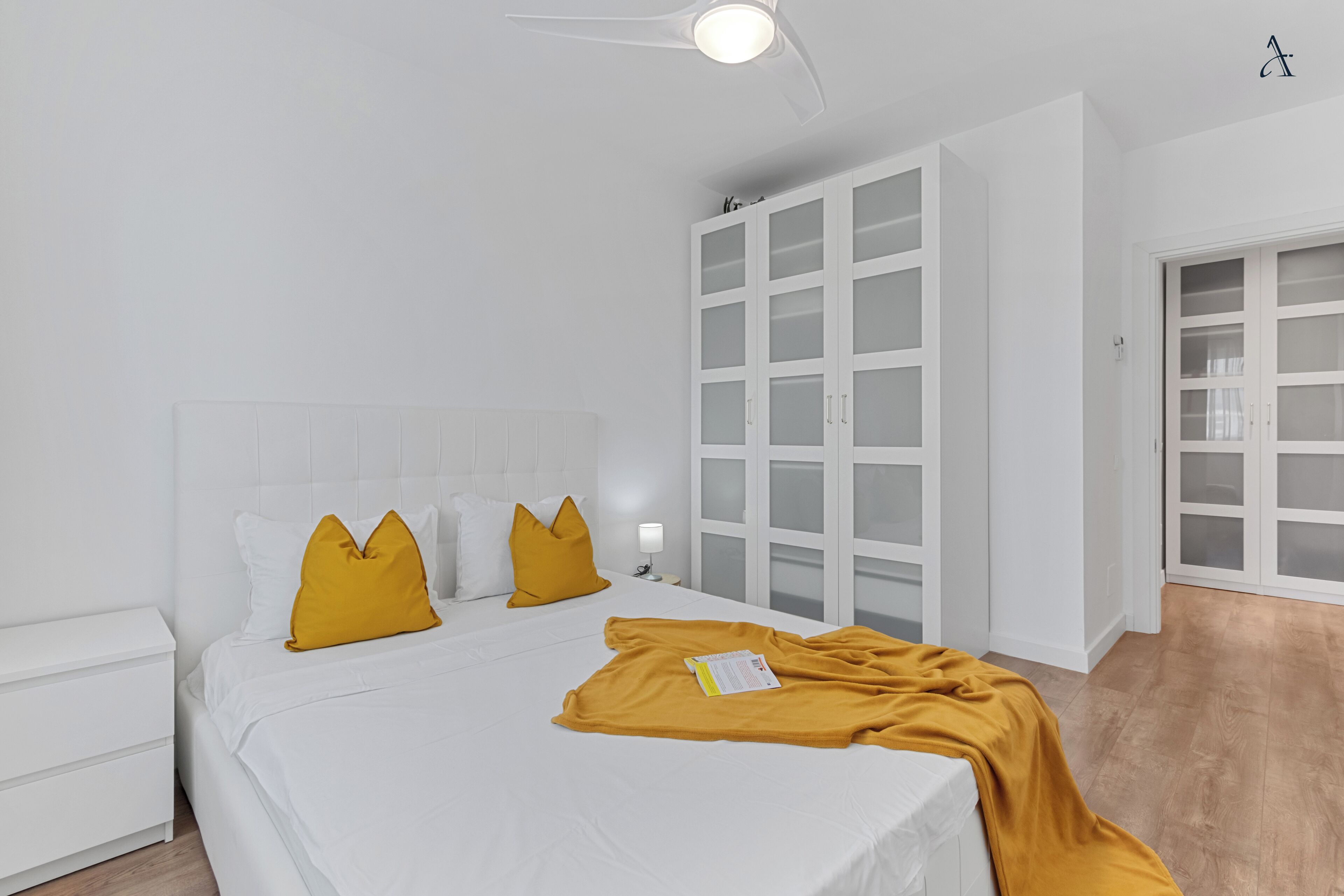 A bedroom with a white bed, mustard-colored pillows, a white nightstand, a white wardrobe, and a ceiling fan.