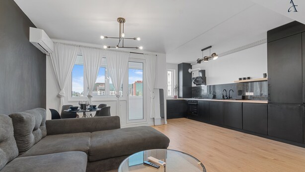 Un apartamento moderno con un sofá gris, una mesa de centro de cristal, una cocina con armarios negros y vistas de la ciudad a través de amplias ventanas.