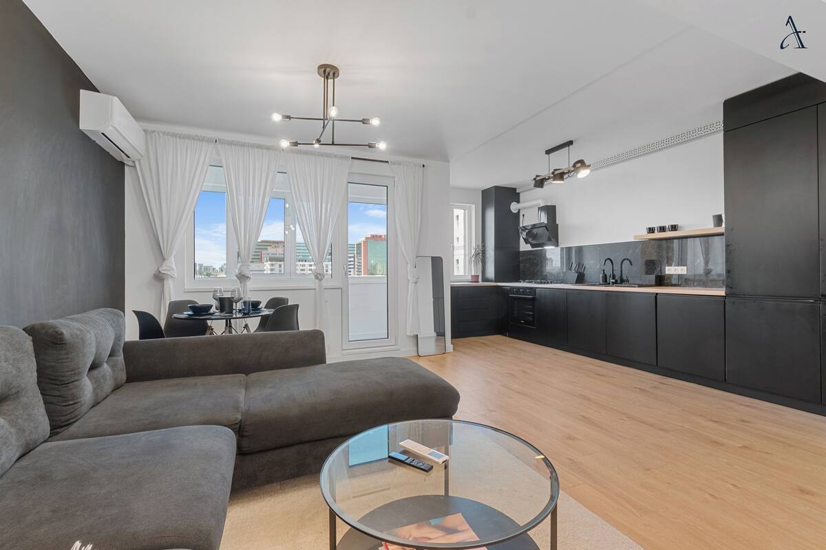 Eine moderne Wohnung mit grauem Sofa, einem Glastisch, einer Küche mit schwarzen Schränken und Blick auf die Stadt durch große Fenster.