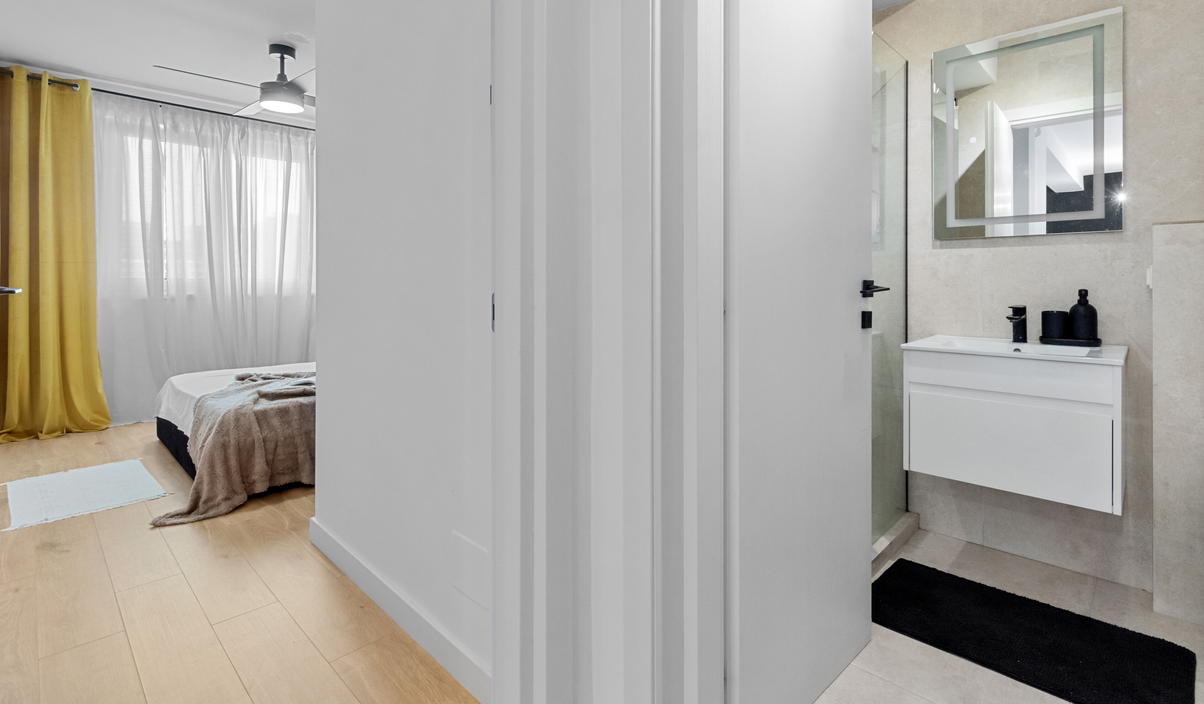 A modern hotel room with a bed, a bedside table, a mirror, and a bathroom with a sink and mirror.