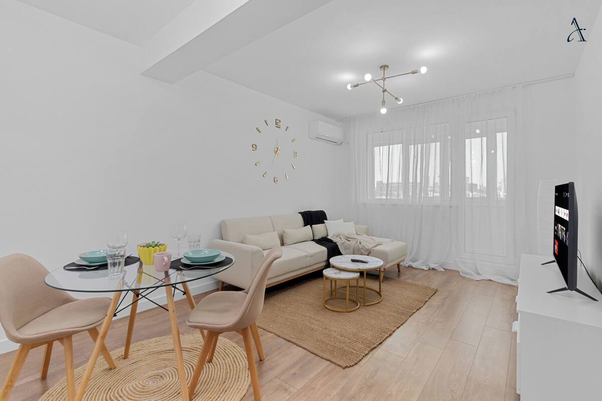 Ein modernes Wohnzimmer mit einem Glastisch, einem beigefarbenen Sofa und einem Fernseher auf einem weißen Schrank.