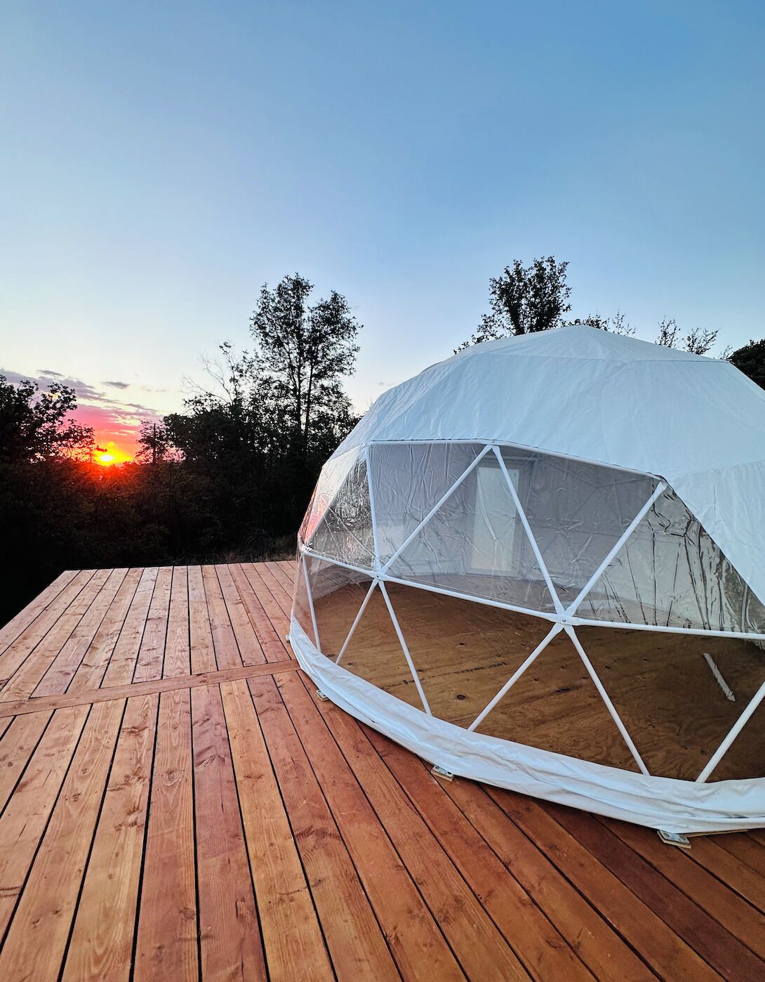 Ein Geodätik-Dome-Zelt auf einer Holzterrasse, im Hintergrund ein Sonnenuntergang.