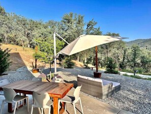 Eine Terrasse im Freien mit Holztisch und -stühlen, einem großen Sonnenschirm und Blick auf einen bewaldeten Hang.