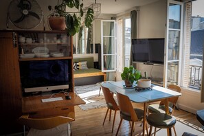 A cozy living room with a wooden desk, a large flat-screen TV, and a comfortable seating area with a sofa and a dining table. - Charmant Appartement Haut-marais Paris (Paris)