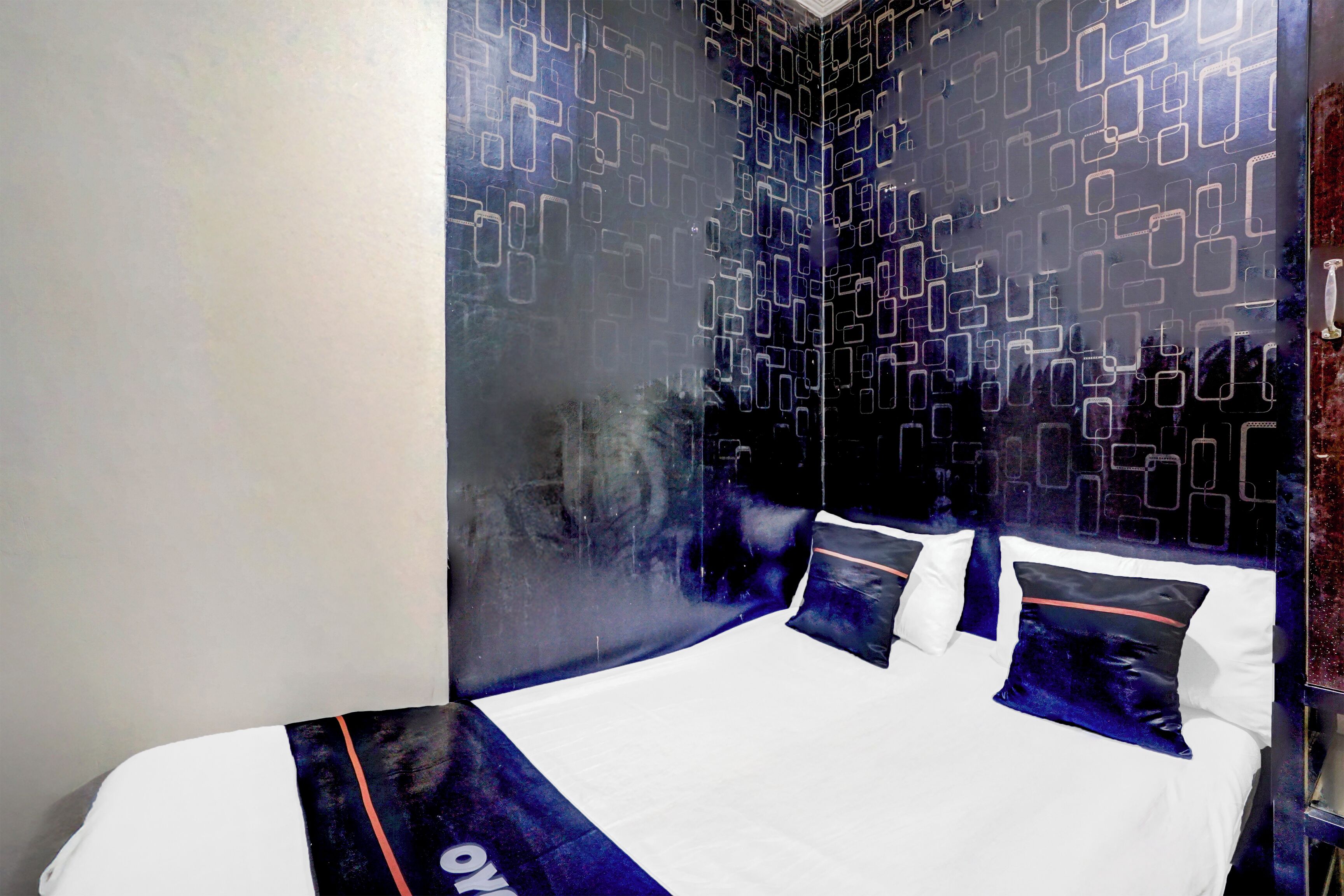 A modern hotel room with a geometric patterned wall, a neatly made bed with white and dark blue bedding, and a visible logo on the bedspread.