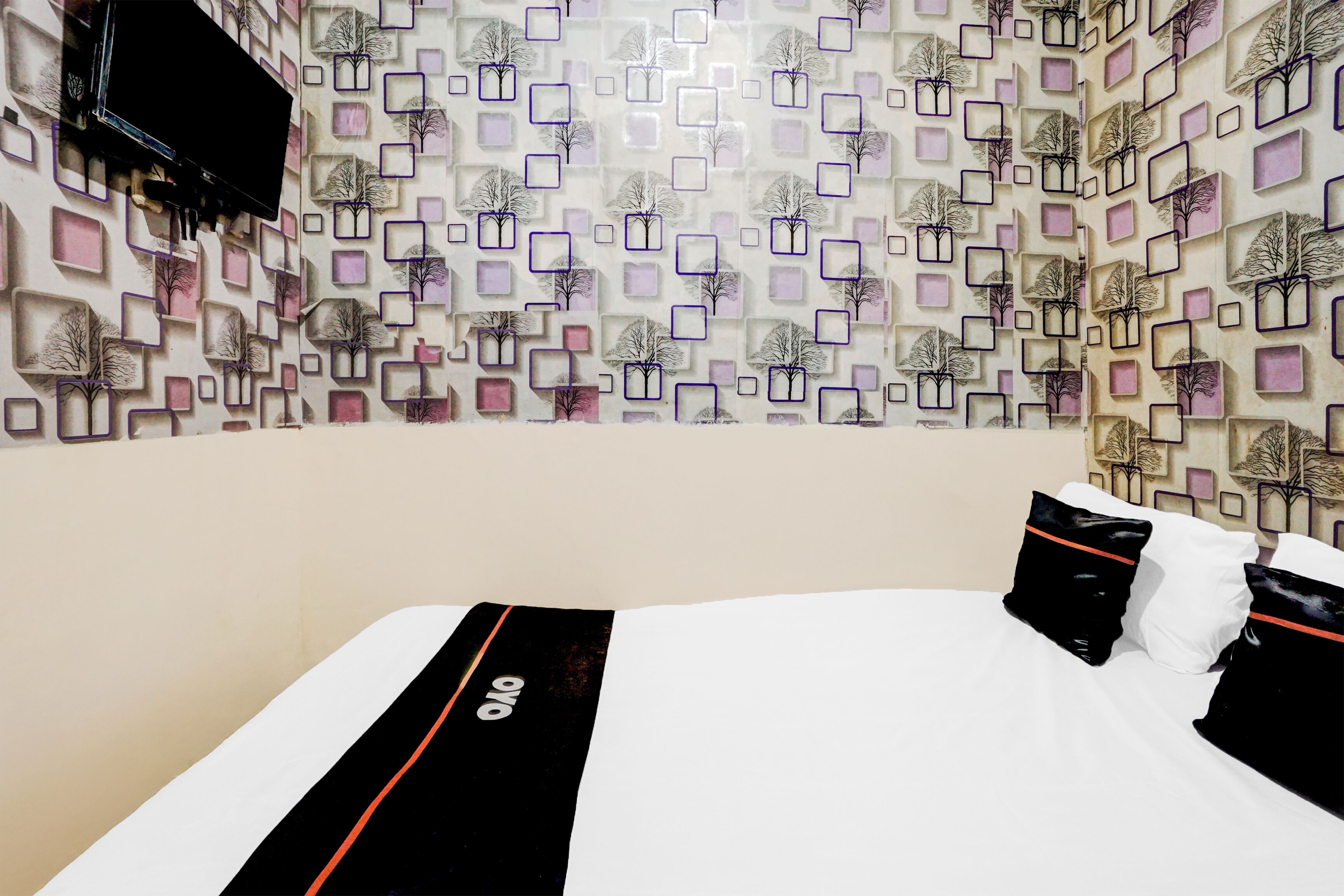 A hotel room with a patterned wall, a bed with white bedding and black pillows, and a flat-screen TV mounted on the wall.