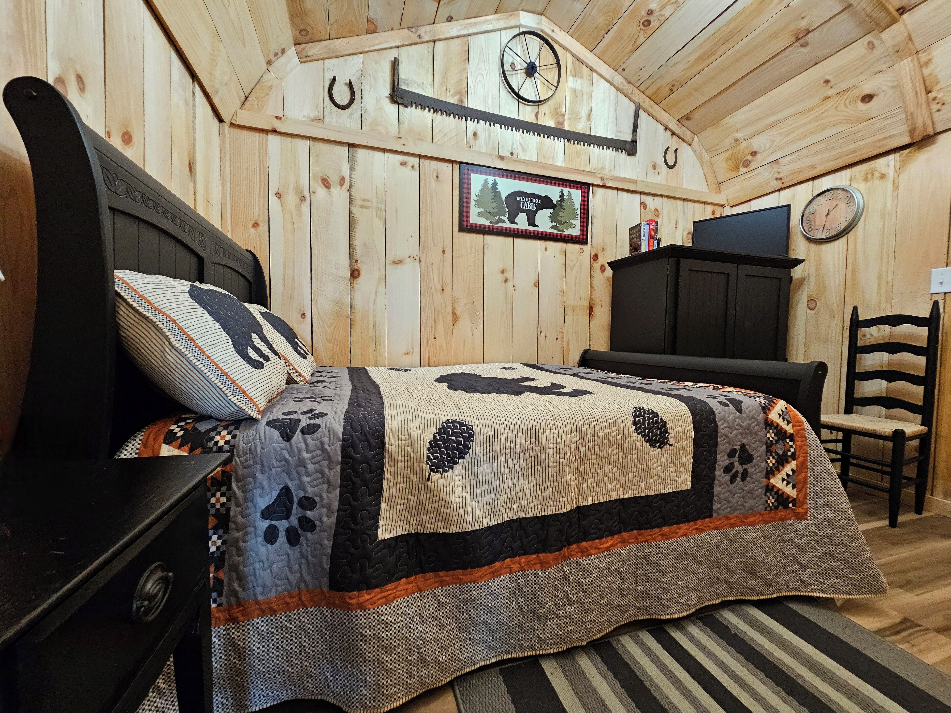 Une chambre à coucher dans une cabane en bois, avec un lit, une chaise, une commode, une horloge et un panneau d’accueil encadré.