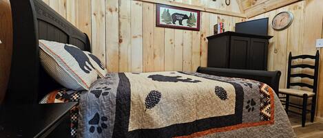 Une chambre à coucher dans une cabane en bois, avec un lit, une chaise, une commode, une horloge et un panneau d’accueil encadré.