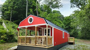 Una cabaña roja con terraza de madera y porche cubierto, rodeada por un camino de grava y abundante vegetación.