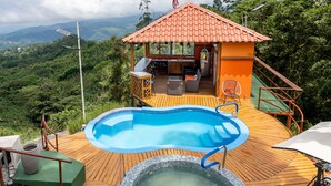 Una terraza de madera con piscina, jacuzzi y un pequeño pabellón con techo rojo.