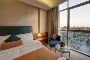 A hotel room with a large bed, a small table, a chair, and a view of the city through large windows.