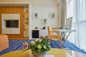 A hotel room with a dining table, a small kitchen area, a television, and a decorative vase with flowers.