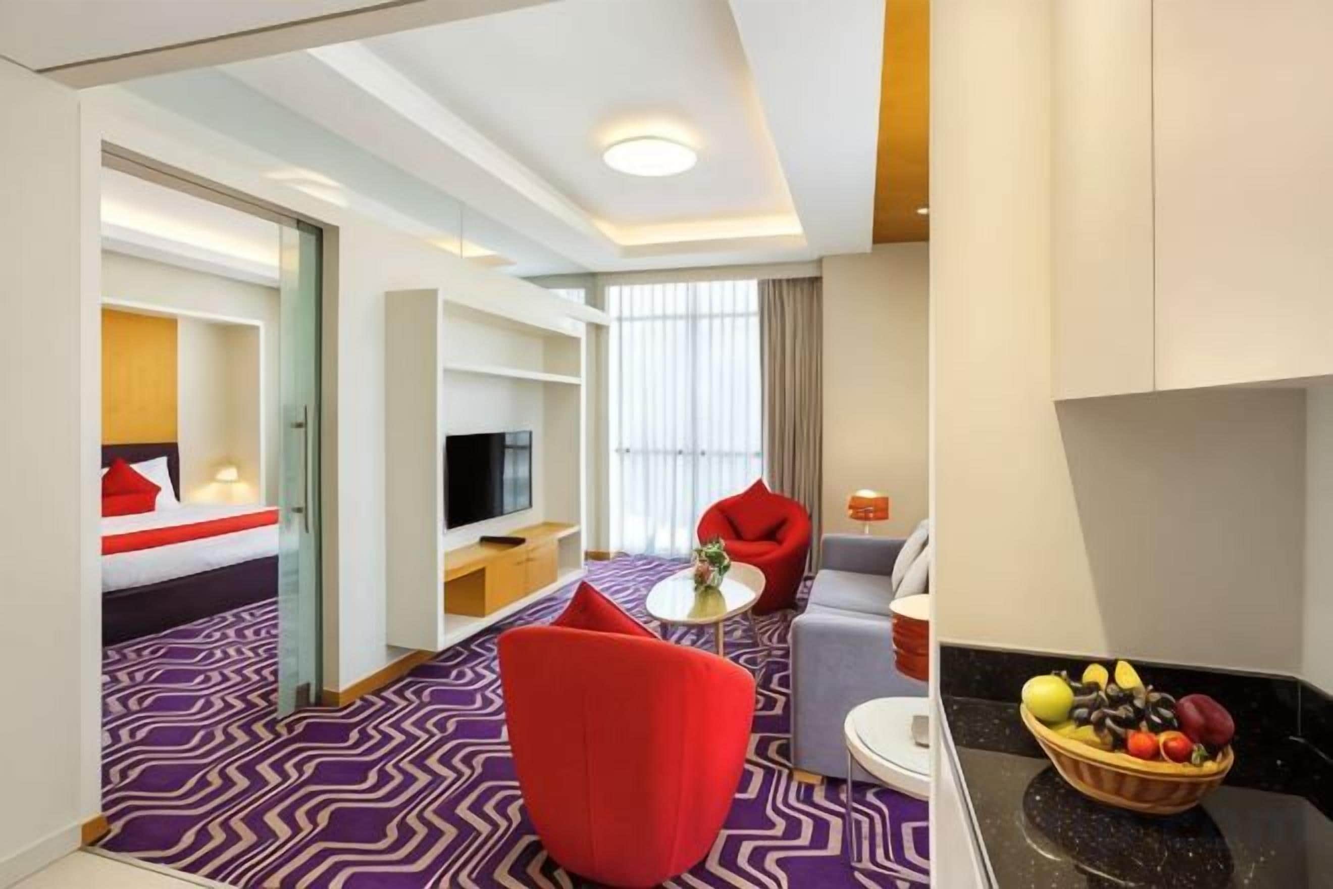 Habitación de hotel con cama, televisor, zona de comedor con mesa y sillas, y frutero.