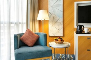 Een hotelkamer met een blauwe fauteuil, een ronde tafel met een schaal fruit, een lamp en een televisie aan een houten wand.