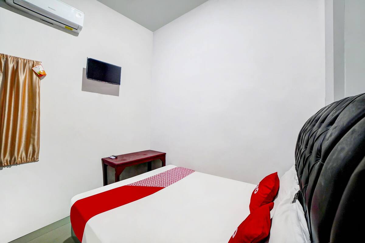 A hotel room with a bed, a small table, a wall-mounted TV, and an air conditioning unit.