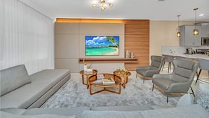 A modern living room with a sofa, coffee table, and chairs, a flat-screen TV, and a kitchen area. - Storey Lake 9 BR Themed Games Room (Kissimmee)