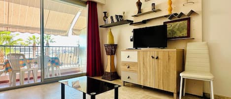 A living room with a glass door leading to a balcony, a flat-screen TV, a cabinet with drawers, a white chair, and a small table.