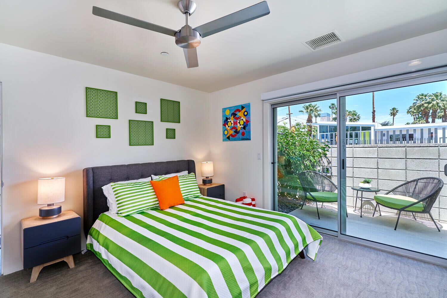 A modern bedroom with a large bed, two bedside tables, a ceiling fan, and a sliding glass door leading to an outdoor area with chairs and a table.