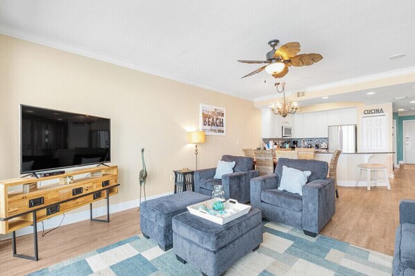 Sala de estar con televisor de pantalla plana, sofás grises, mueble de entretenimiento de madera, ventilador de techo y área de cocina al fondo.