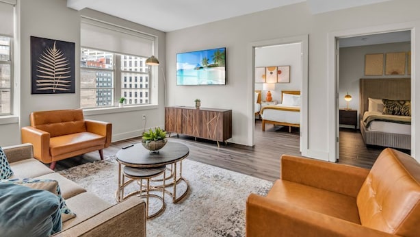A modern living room with a sofa, a coffee table, and a TV. There is a bed in the background and a window with a view of buildings. - Luxury 4BD Condo Near French Quarter – Prime Location! (New Orleans)