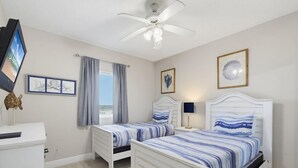 Ein Schlafzimmer mit zwei Betten, einem Deckenventilator, einem Fernseher und einem Fenster mit Vorhängen.