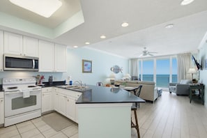 A modern kitchen with white cabinets, a black countertop, and a large island. There is a dining area with a sofa and chairs, and a balcony with a view of the ocean.