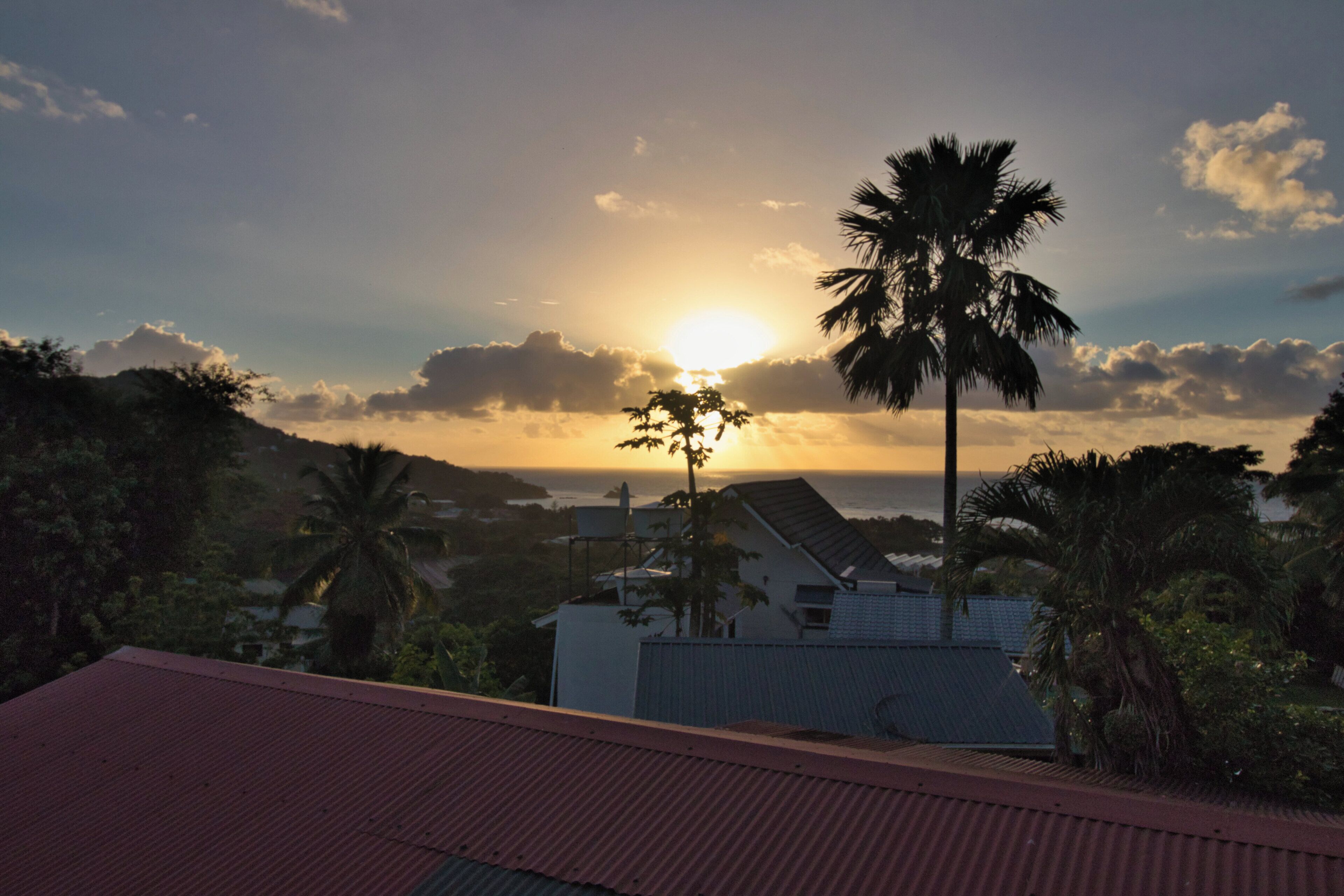 Une vue sur le coucher de soleil, prise depuis un bâtiment avec un toit rouge, des palmiers et une étendue d'eau au loin.