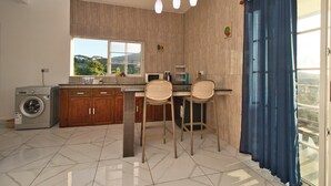 A kitchen with a washing machine, wooden cabinets, a window with a view, and two bar stools. - Creole Cottage 2BR Apartment is a fully furnished located in Anse Royal with AC (victoria)