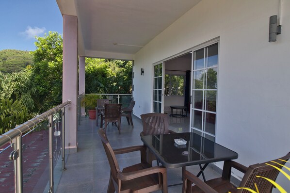 A balcony with a glass table and chairs, a potted plant, and a view of greenery.