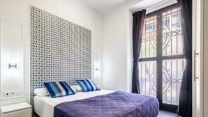 Ein Schlafzimmer mit einem Bett, einem gemusterten Kopfteil, einem Fenster mit Vorhängen und einem weißen Nachttisch.