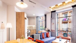 A modern living room with a wooden accent wall, a dining table with chairs, a sofa with colorful cushions, and a view of the street through large windows. - Beautiful Suite with Private Terrace (Valencia)