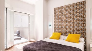 Quarto com cama, dois travesseiros amarelos, parede estampada e porta de vidro deslizante para uma varanda.