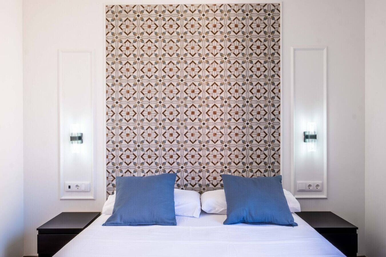 A hotel room with a patterned headboard, two bedside tables, and two blue pillows.