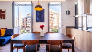 A modern dining area with a wooden table and chairs, a vase with a red flower, and a view of buildings through large windows. - Well Lit and Creatively Designed Apartment (Valencia)