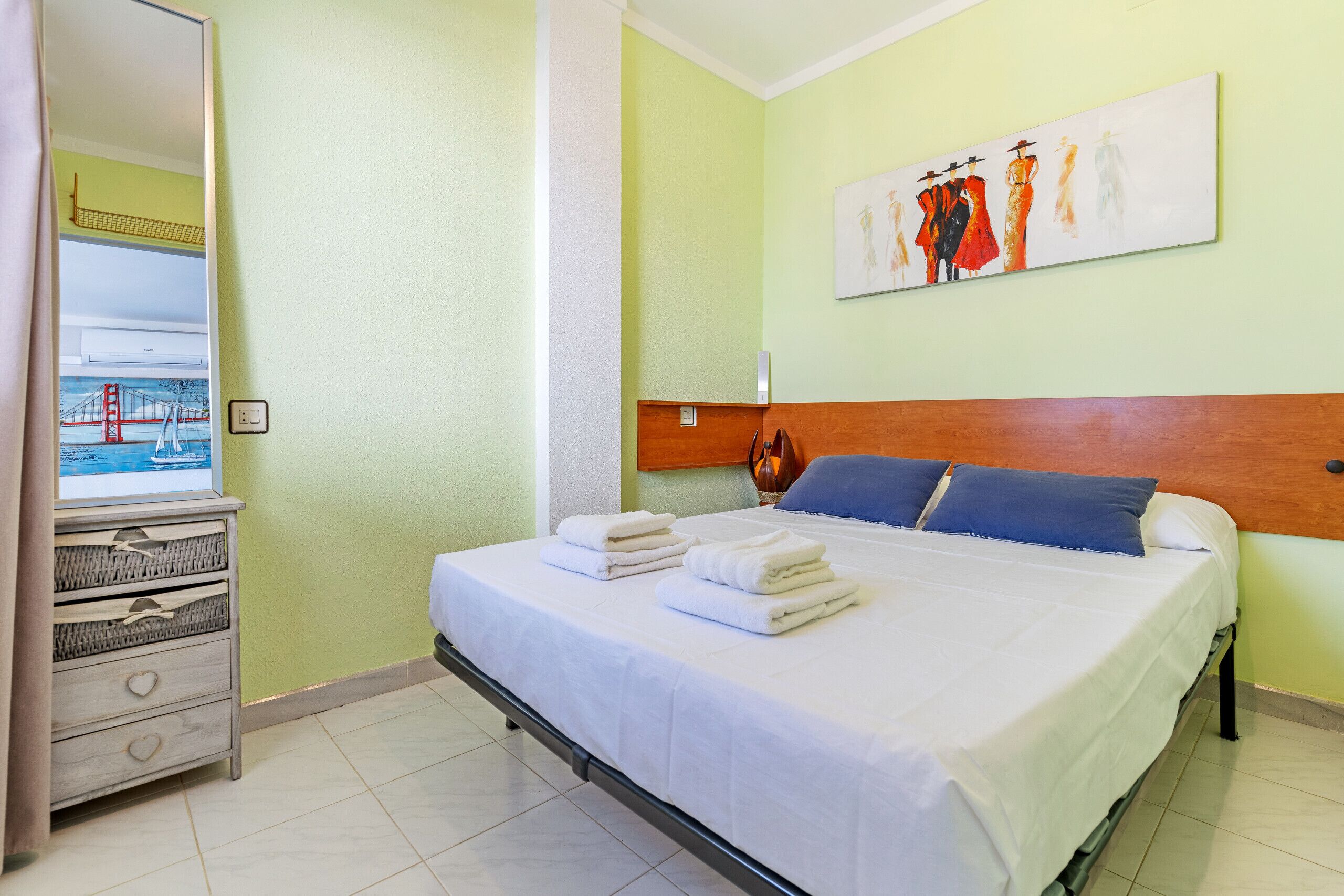 Una cama bien hecha con sábanas blancas y almohadas azules, un aparador con cajones, un cuadro en la pared y una ventana con vistas a un puente.