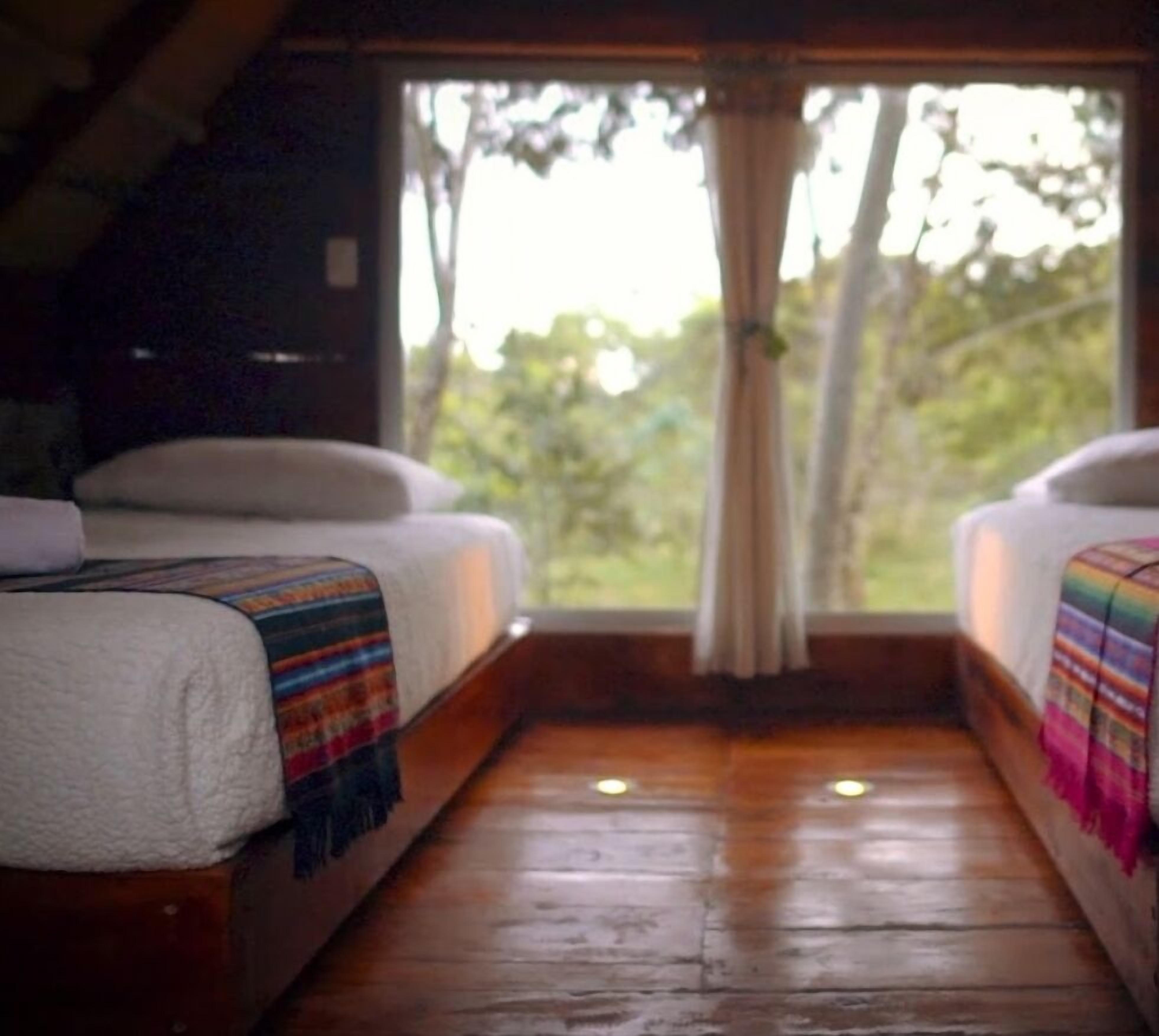 A room with two beds, each with a colorful blanket, wooden flooring, and a window with curtains.
