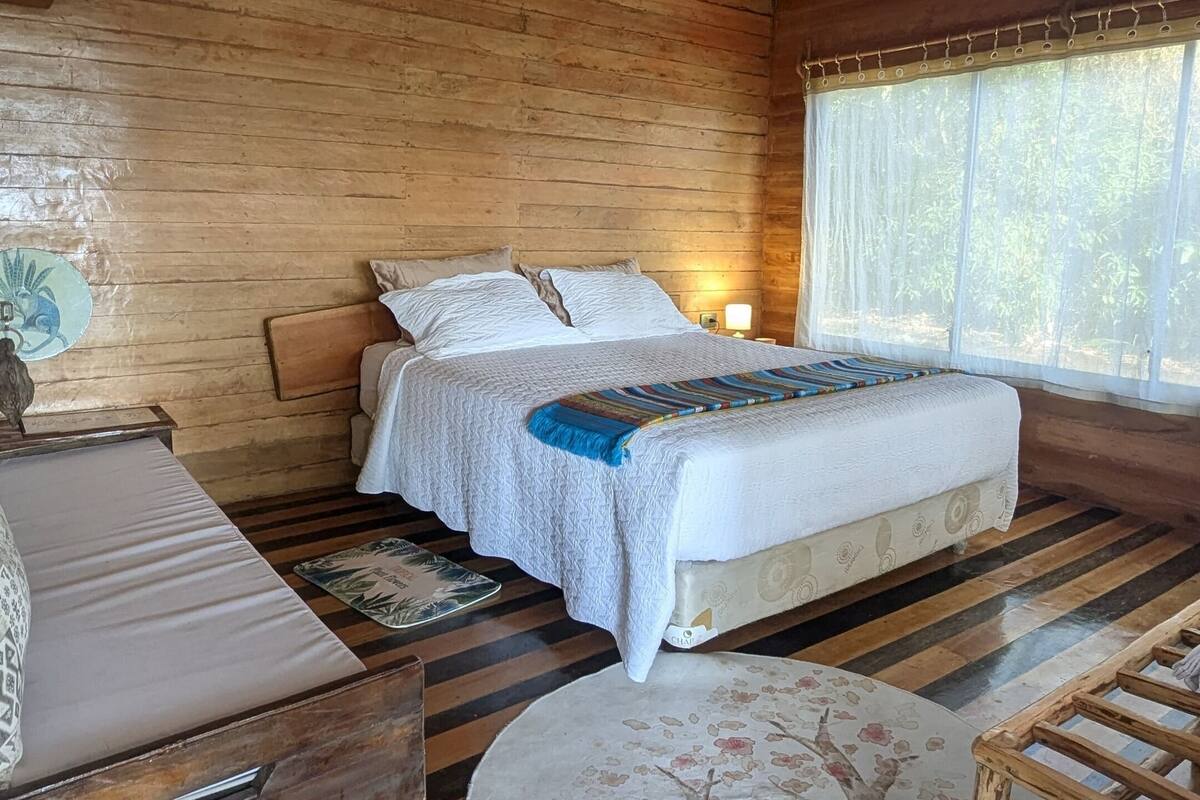 Une chambre dans une cabane en bois, avec un lit, une chaise, une table et une fenêtre avec des rideaux.