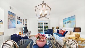 A modern living room with a glass coffee table, blue and white sofas, a pink vase with flowers, and artwork on the walls.