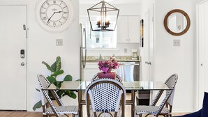A dining area with a glass table, wicker chairs, a wall clock, and a hanging light fixture.