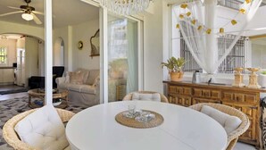 A spacious living area with a round white table, wicker chairs, a chandelier, and a view through a glass door to a kitchen and dining area. - Garden Pool Apartment in Mijas by Costarentals - Ref 209 (Las Lagunas de Mijas)