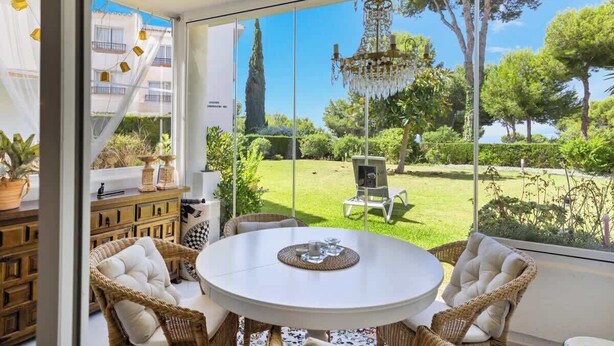 A sunlit room with a round table, wicker chairs, and a view of a lush garden through large windows. - Garden Pool Apartment in Mijas by Costarentals - Ref 209 (Las Lagunas de Mijas)