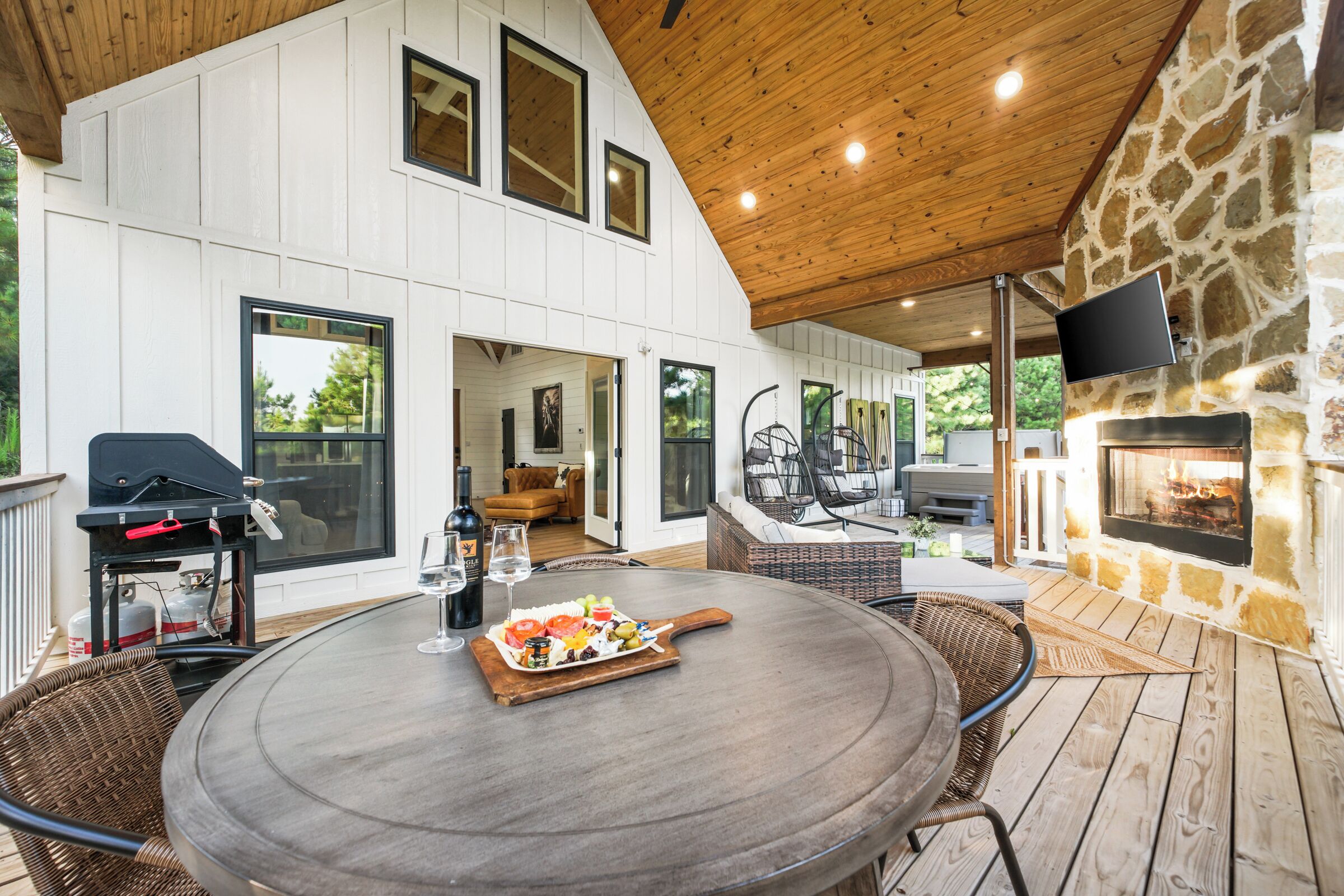 Een overdekt buitenruimte met een ronde tafel, rieten stoelen, een barbecue en een stenen openhaard.