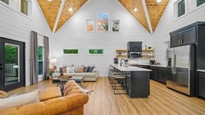 A modern kitchen with a central island, a dining area with a sofa, and a large window overlooking greenery. - Romantic retreat, Hot tub, firepit and games! (Broken Bow)